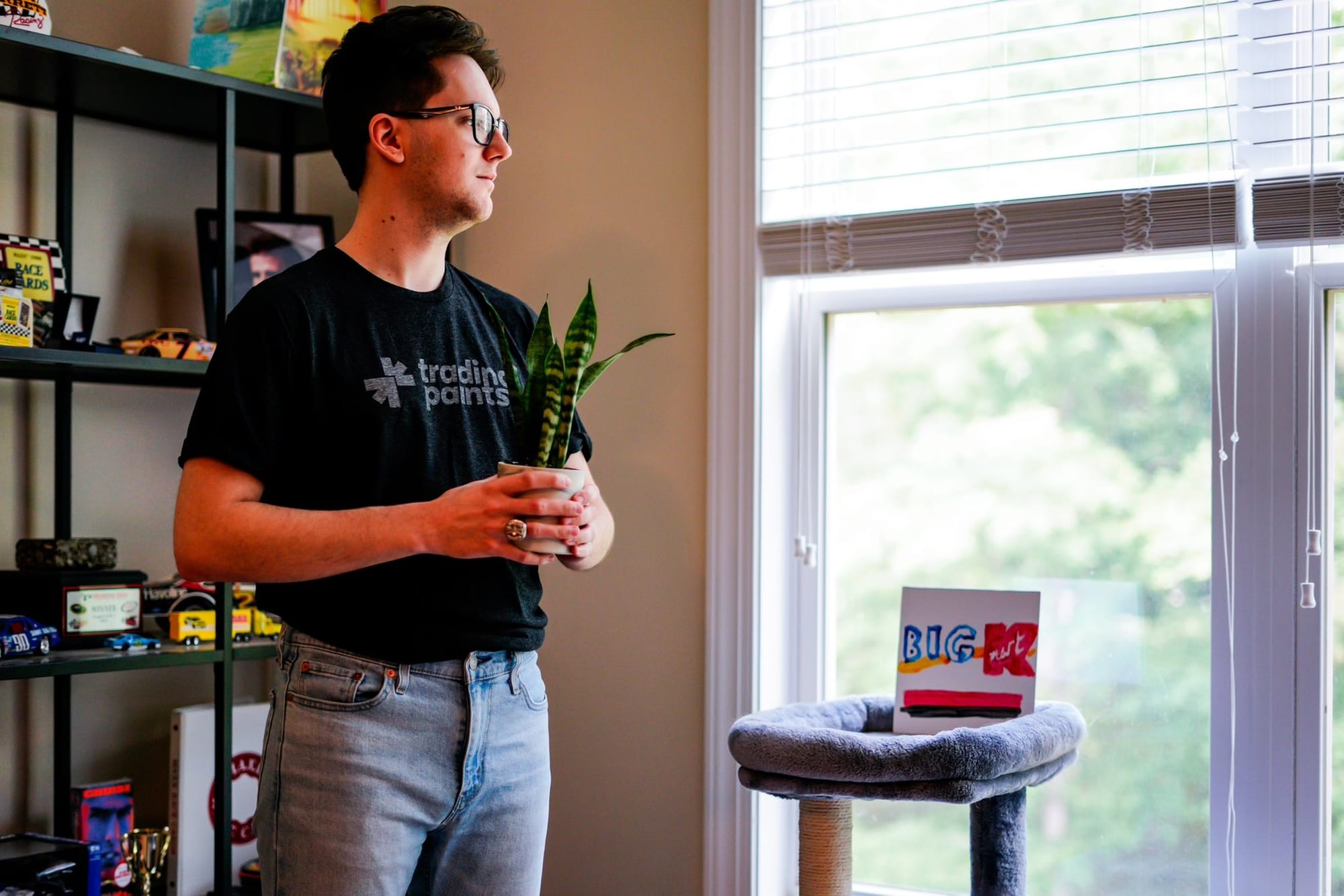 Ryan Williams, wearing a Trading Paints shirt, holding a snake plant, peering longingly out a window